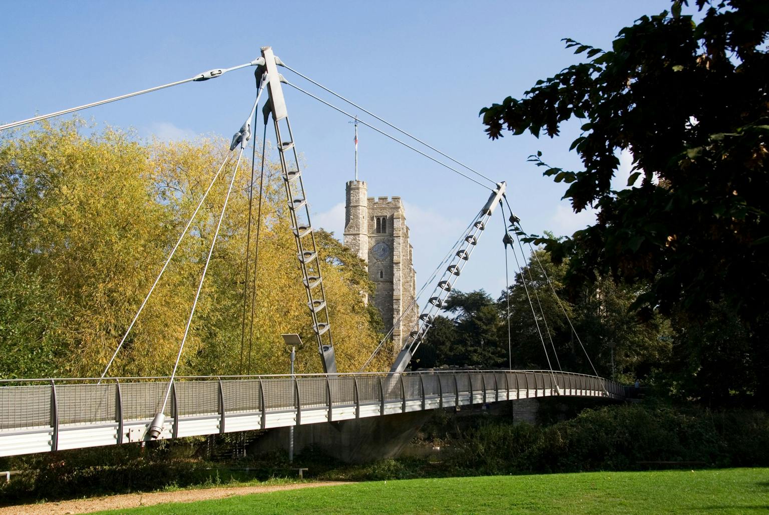 A modern suspension bridge with multiple supports for pedestrians and cyclists crosses the River Medway in Maidstone, Kent, England. In the background is a tall office building