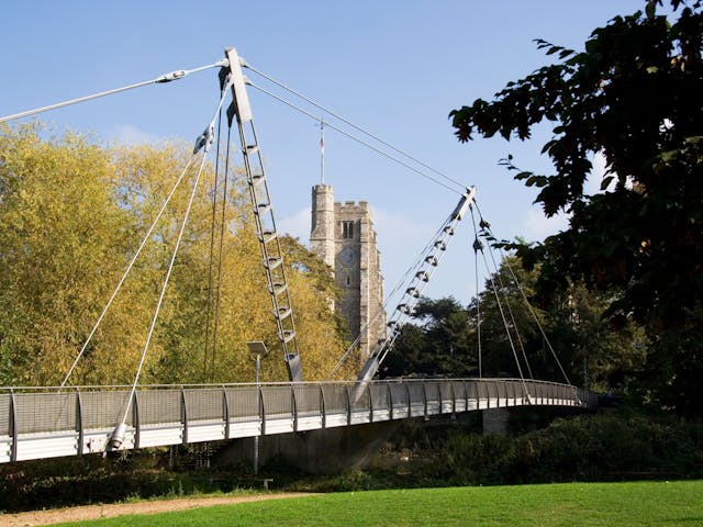 A modern suspension bridge with multiple supports for pedestrians and cyclists crosses the River Medway in Maidstone, Kent, England. In the background is a tall office building