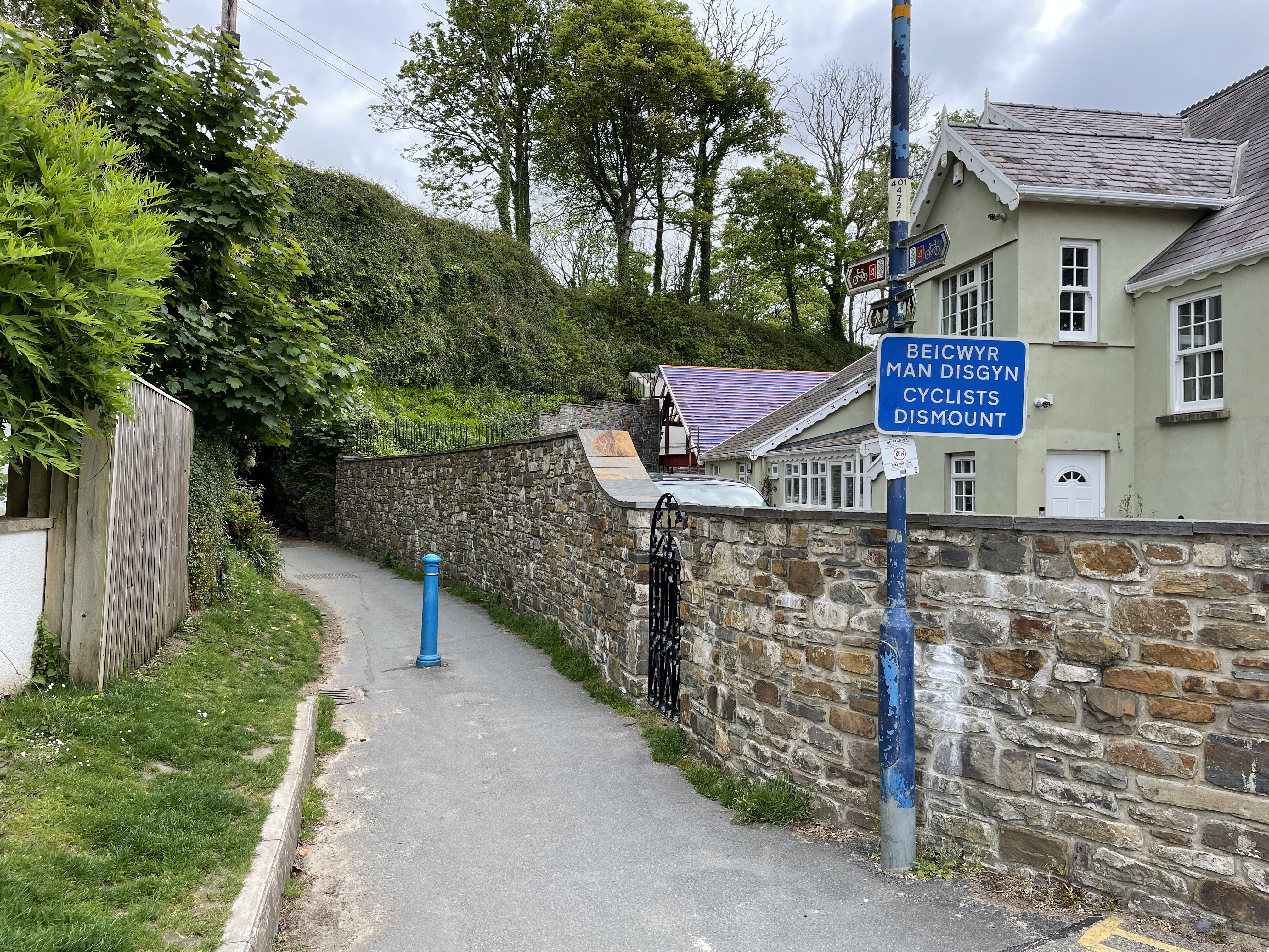 Blue sign on a pole next to a brick wall, with text in Welsh indicating dismount instructions for cyclists