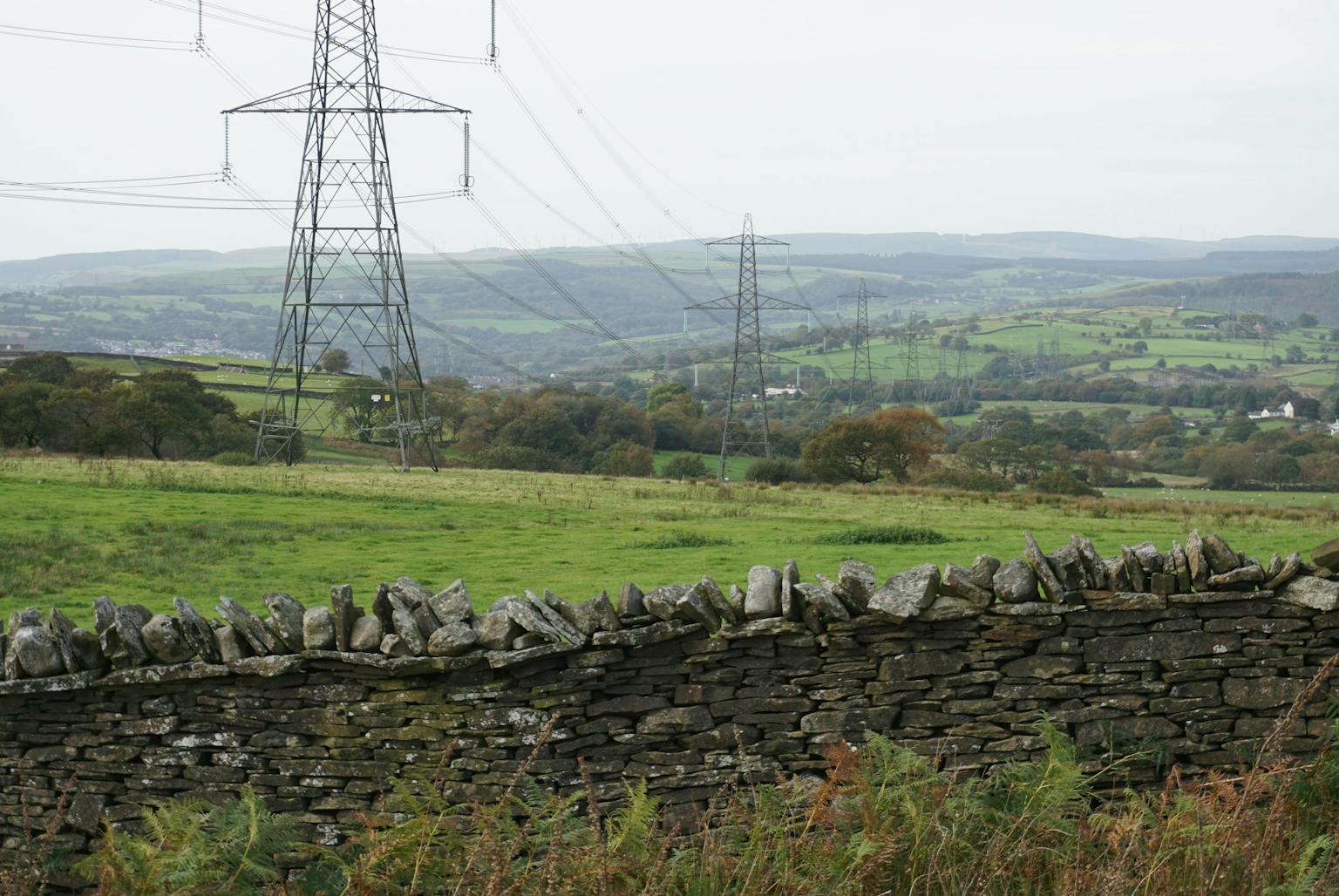 A stone wall made of gray stones separates a grassy field from a rural landscape with power lines in the background