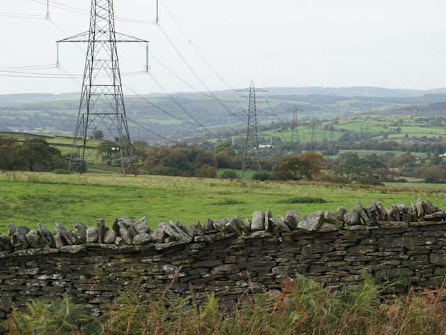 A stone wall made of gray stones separates a grassy field from a rural landscape with power lines in the background