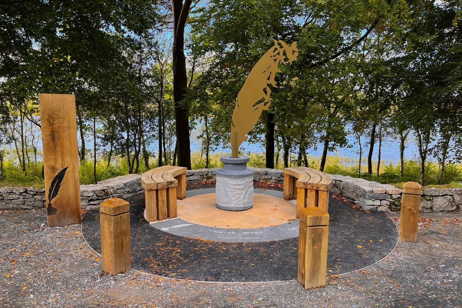 A gold statue of a feather sits on a wooden bench in Yeats Memorial Garden, surrounded by other benches. The garden is located in County Sligo, Ireland