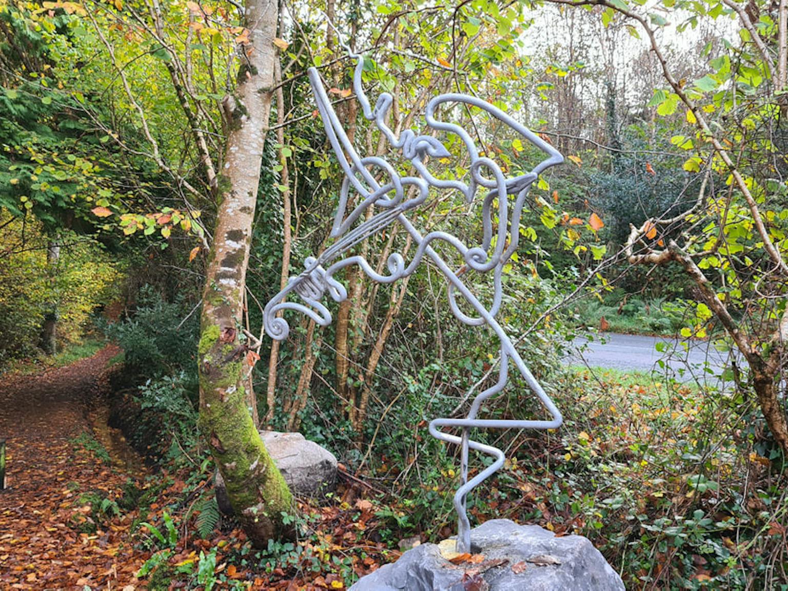 A metal statue of a woman in a flowing dress stands on a stone pedestal in a wooded area. Sunlight filters through the leaves of the trees around the statue.