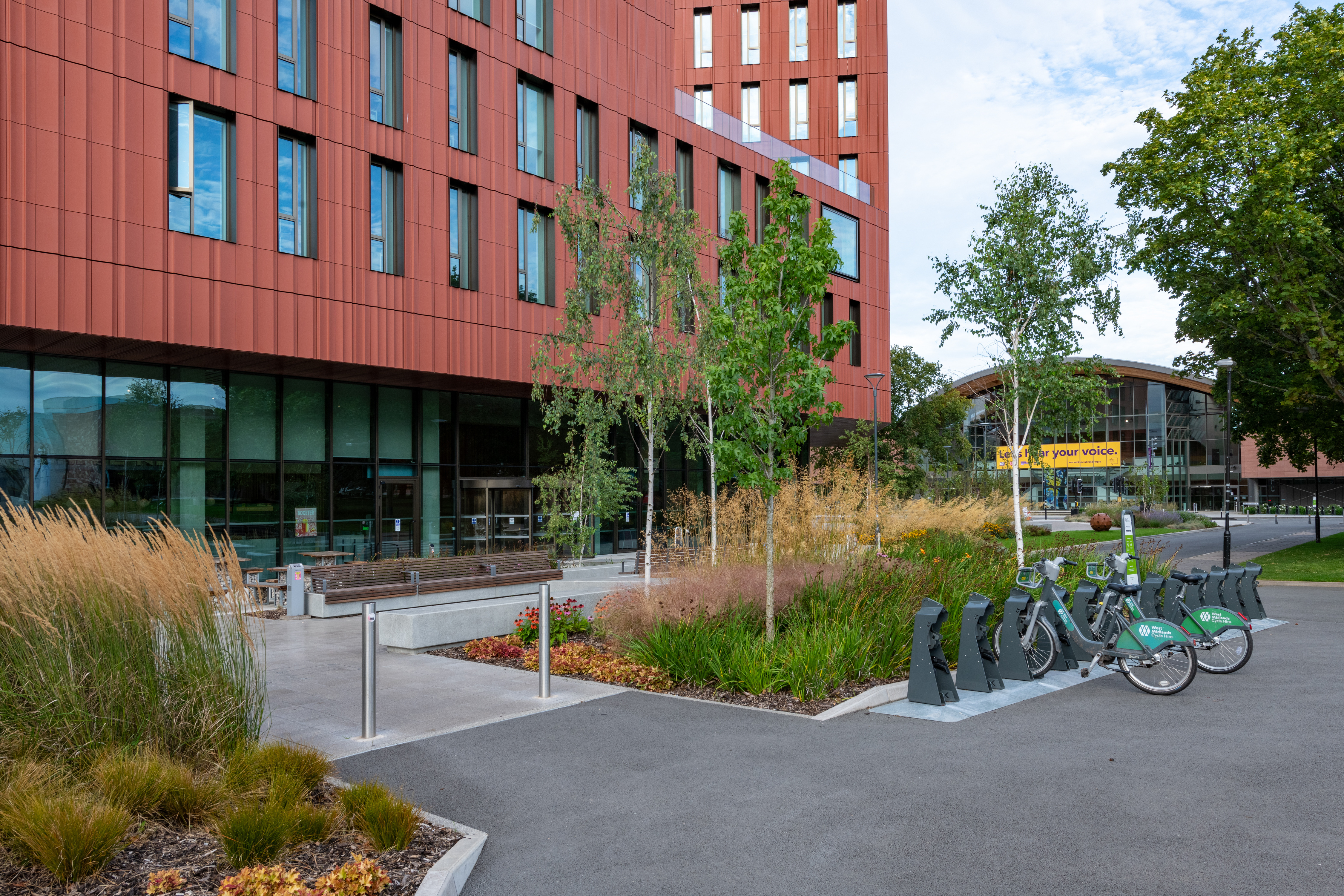 A row of bicycles parked in a rack in front of the University of Warwick's Faculty of Arts building. The building is made of red brick with large windows and decorative terracotta features