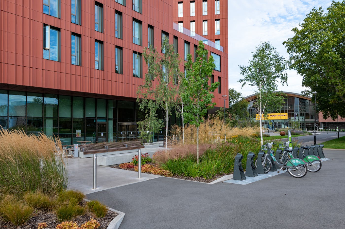 A row of bicycles parked in a rack in front of the University of Warwick's Faculty of Arts building. The building is made of red brick with large windows and decorative terracotta features