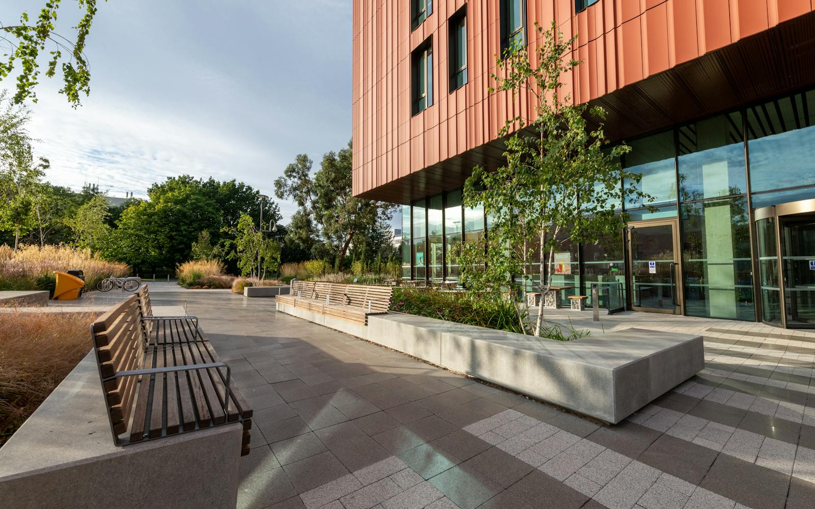 A modern building with a glass facade and a wooden bench with a backrest sits on a sidewalk lined with trees. In front of the bench are planters with bushes