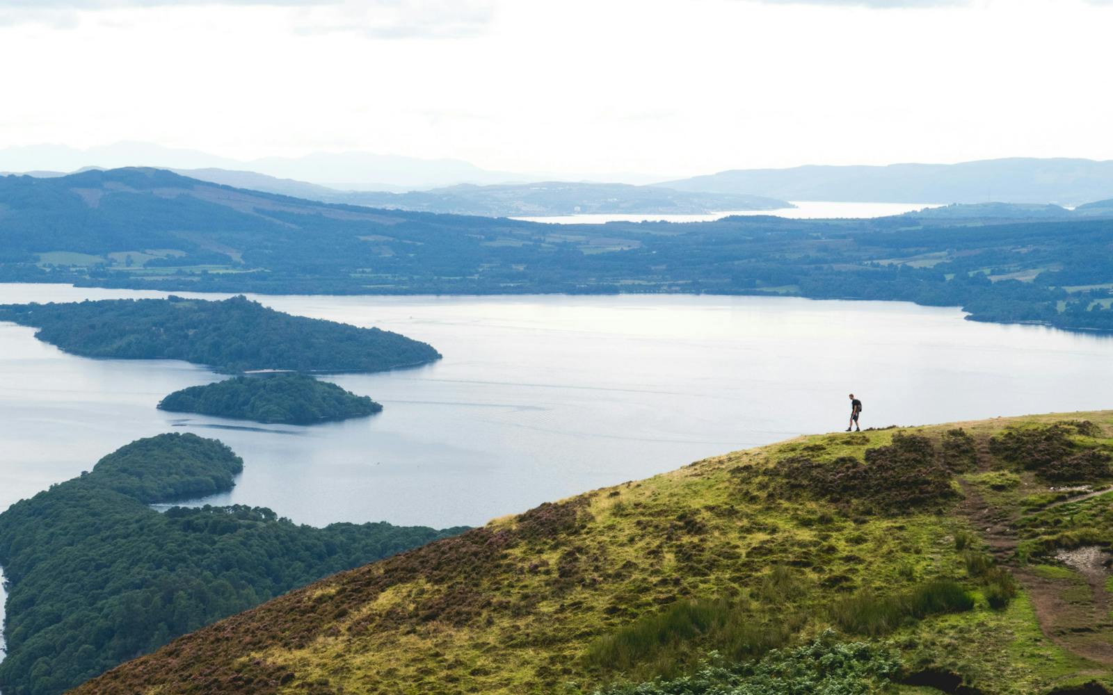 A person standing on a grassy hilltop overlooking a large lake, with mountains in the distance