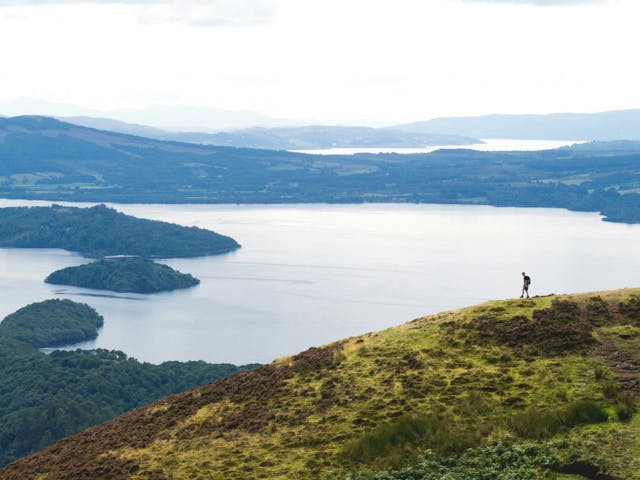 A person standing on a grassy hilltop overlooking a large lake, with mountains in the distance