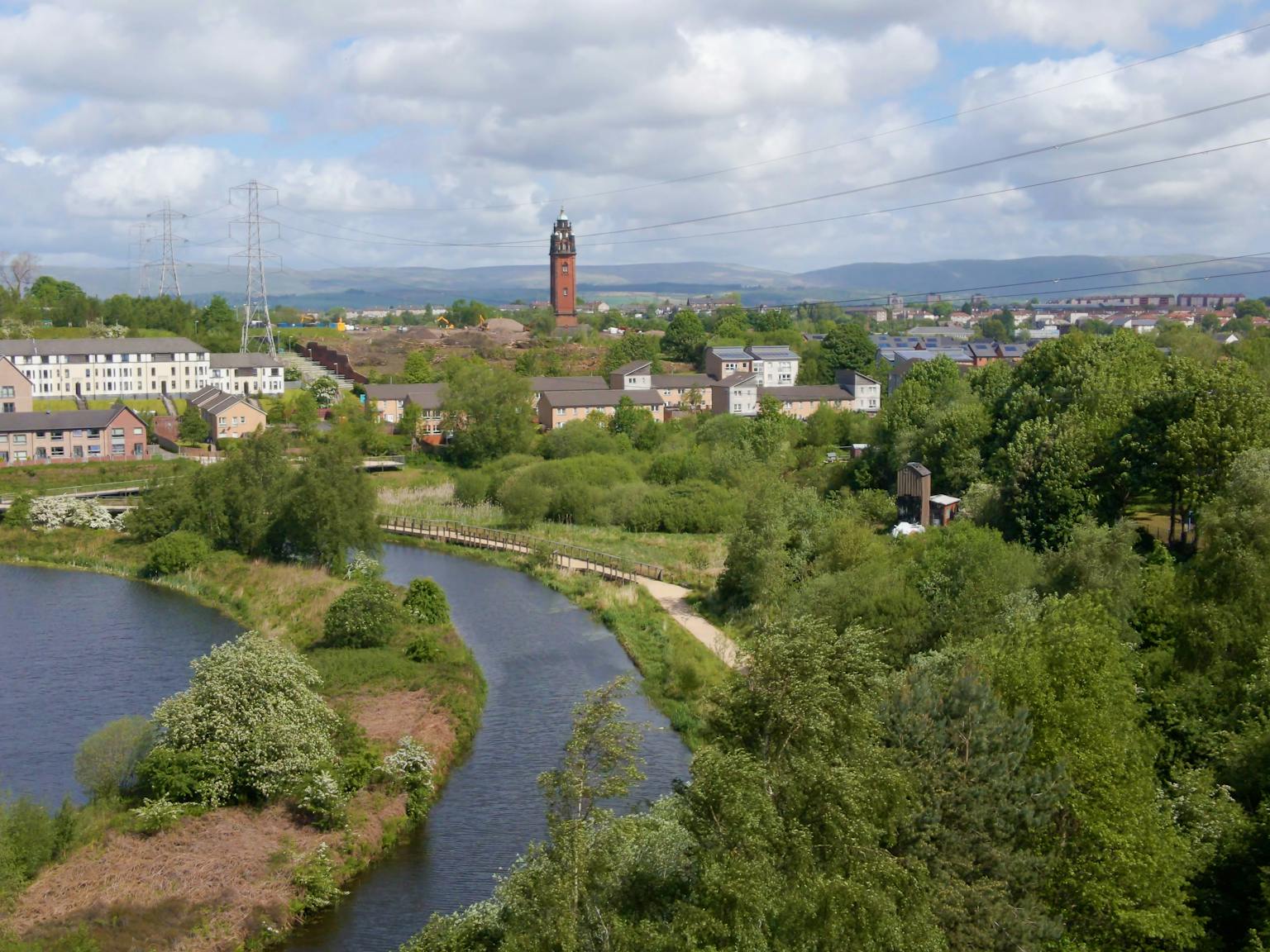 A river and a pond surrounded by trees and greenery overlook a city, with hills in the background