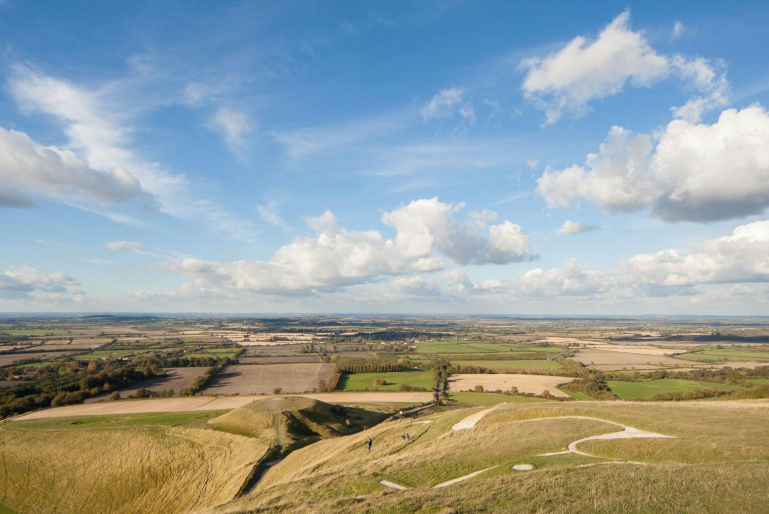 Aerial view of the Uffington White Horse, a prehistoric hill figure carved into the chalk downs of Oxfordshire, England. The horse is surrounded by rolling hills, farmland, and a vast sky with fluffy white clouds