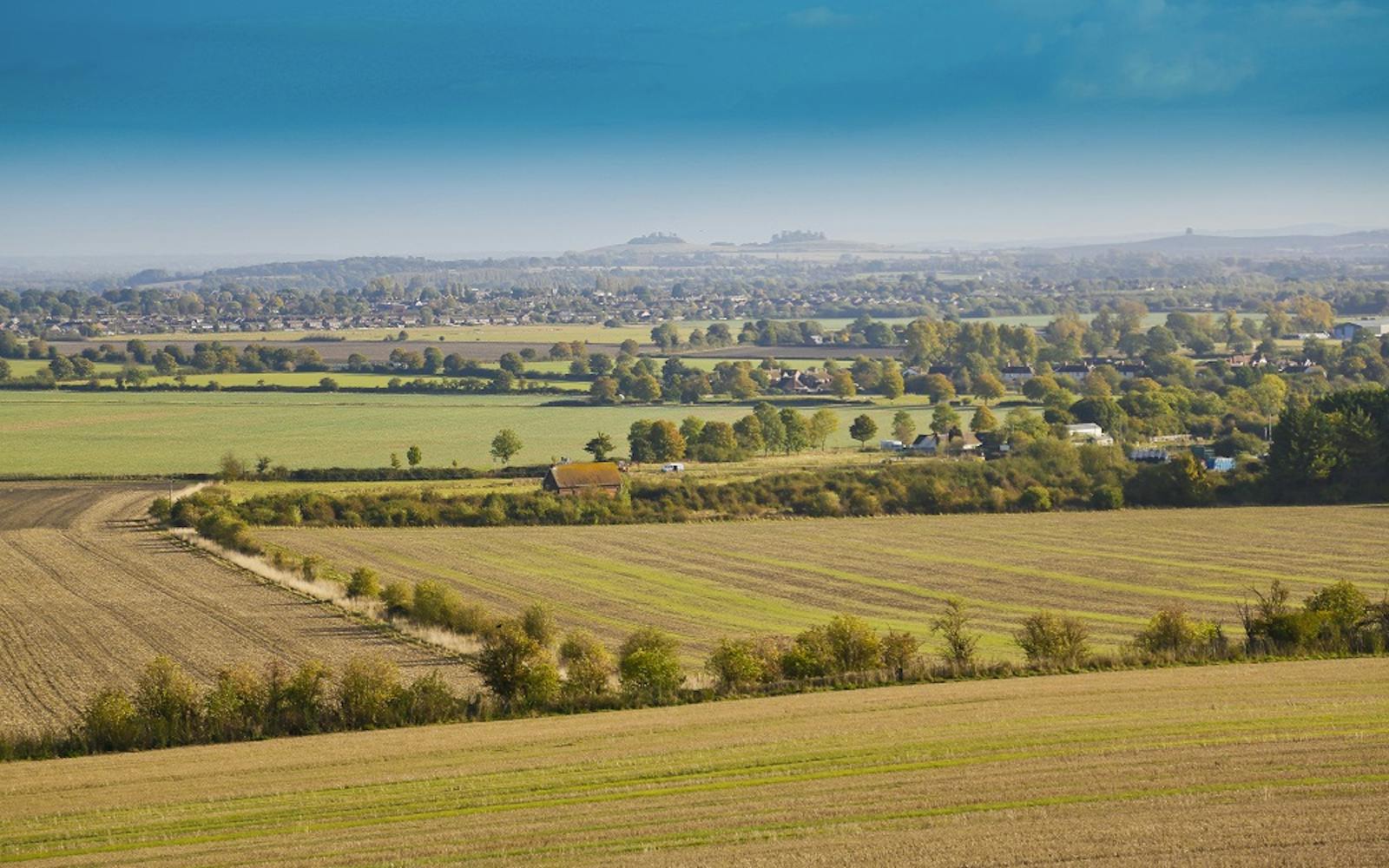 Panoramic view of the Oxfordshire countryside in autumn, featuring rolling fields, scattered trees, farmhouses, and a distant village nestled beneath a blue sky with wispy clouds
