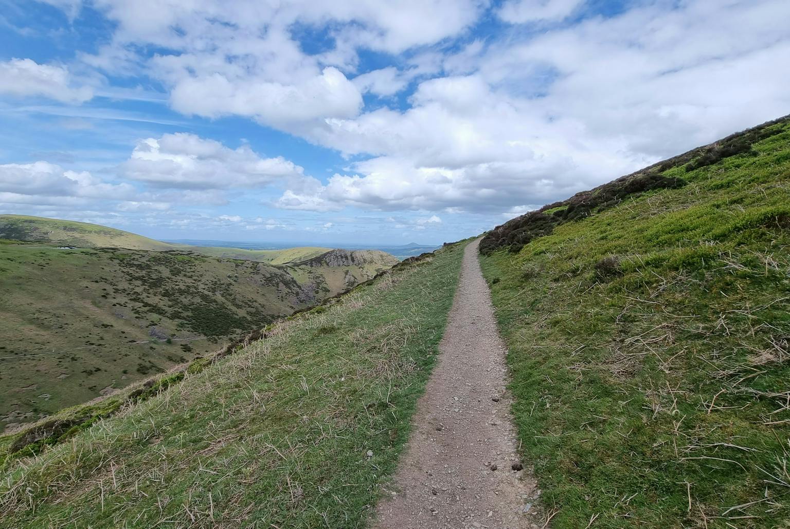 A narrow hiking trail winds along a grassy ridge, overlooking a vast valley under a bright blue sky with fluffy white clouds. Lush green hills and distant peaks create a breathtaking panoramic view
