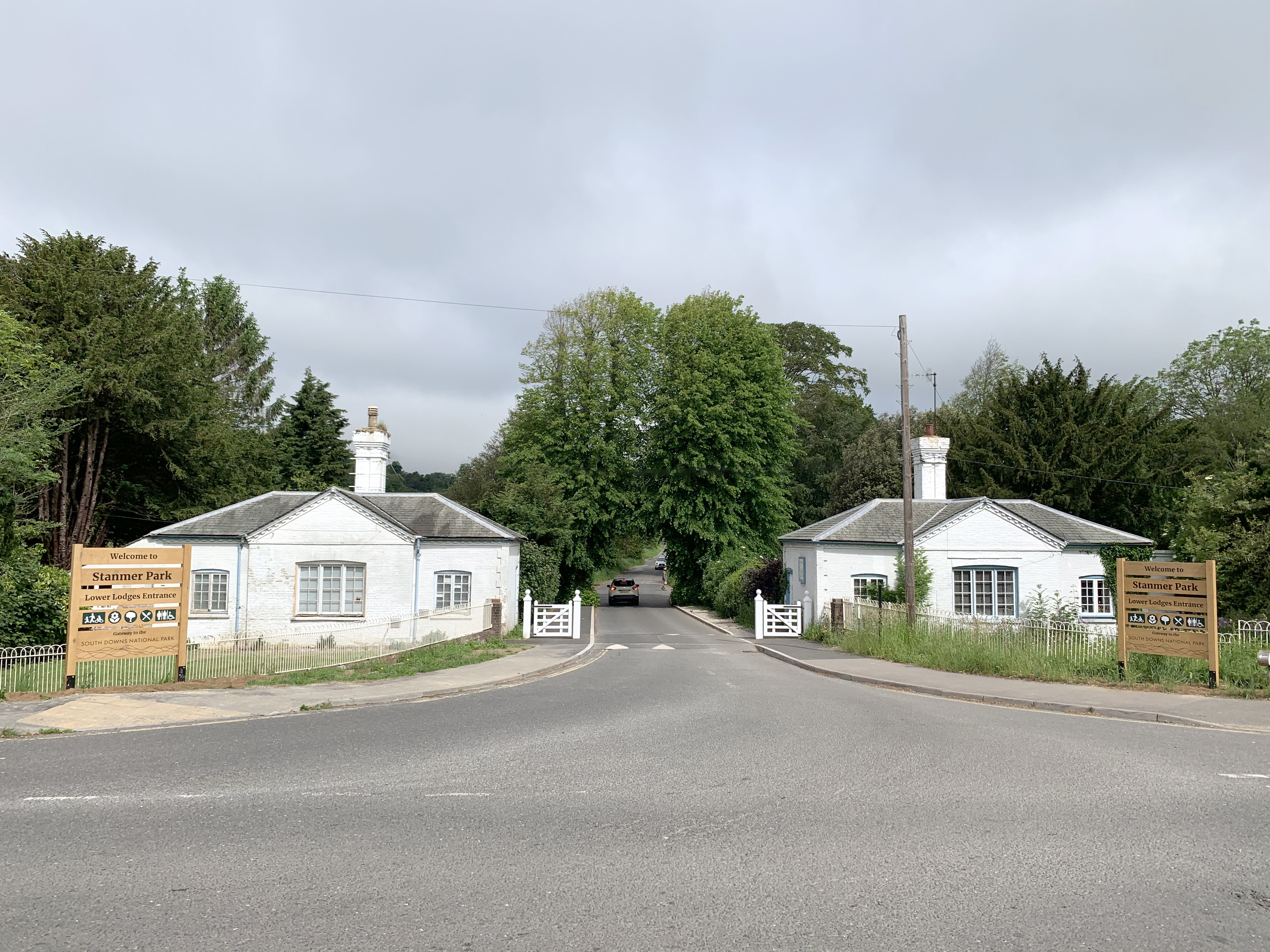 A paved road leads through a gated entrance to Stanmer Park. Two white gatehouses with brown signage flank the entrance. Trees and shrubs line the road beyond the gate.