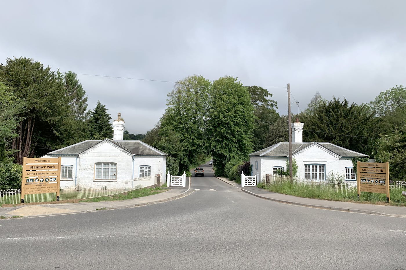 A paved road leads through a gated entrance to Stanmer Park. Two white gatehouses with brown signage flank the entrance. Trees and shrubs line the road beyond the gate.