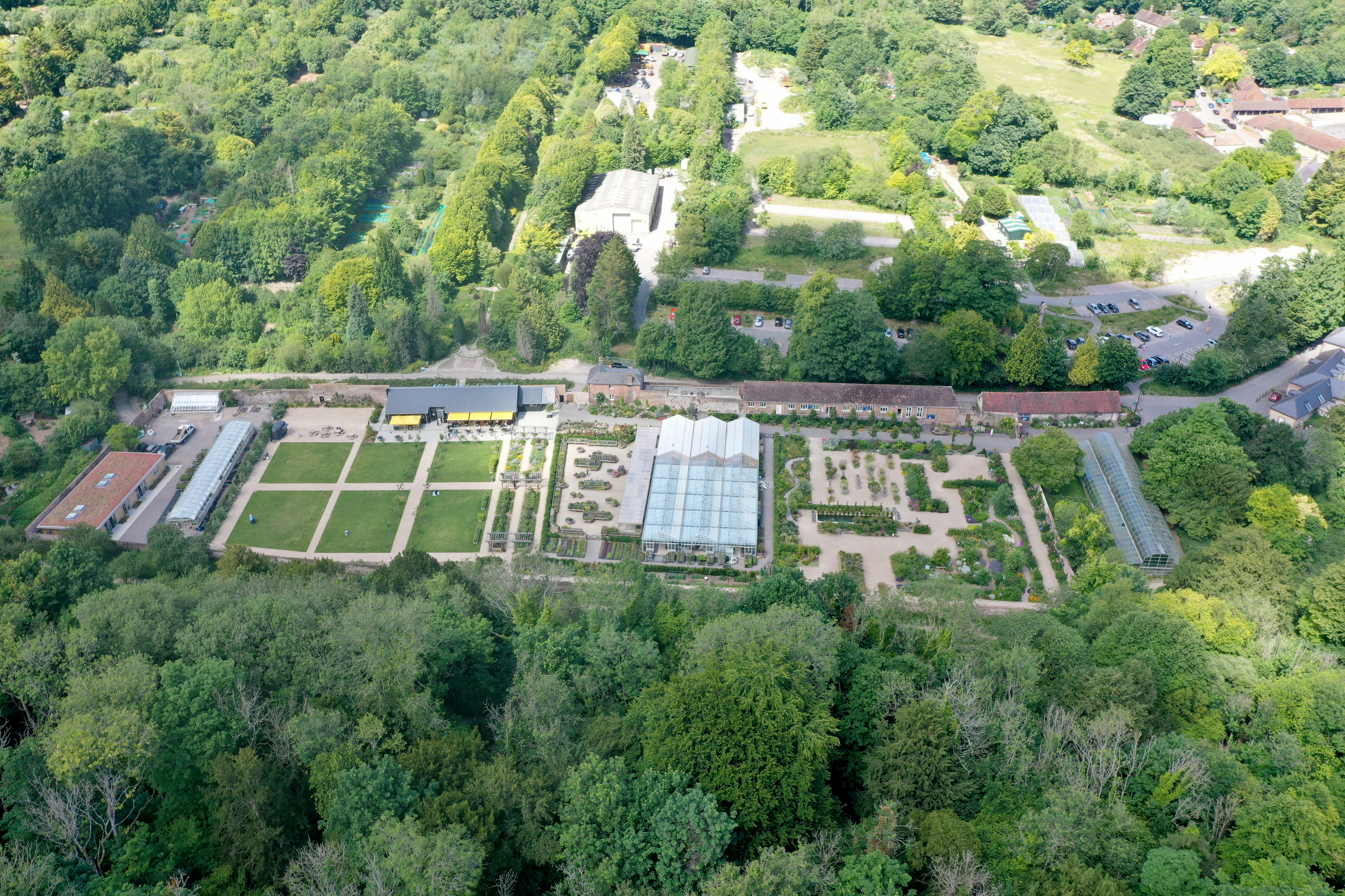 Aerial view of a garden with geometrically arranged garden beds, greenhouses, a visitor centre, and surrounding woodland.