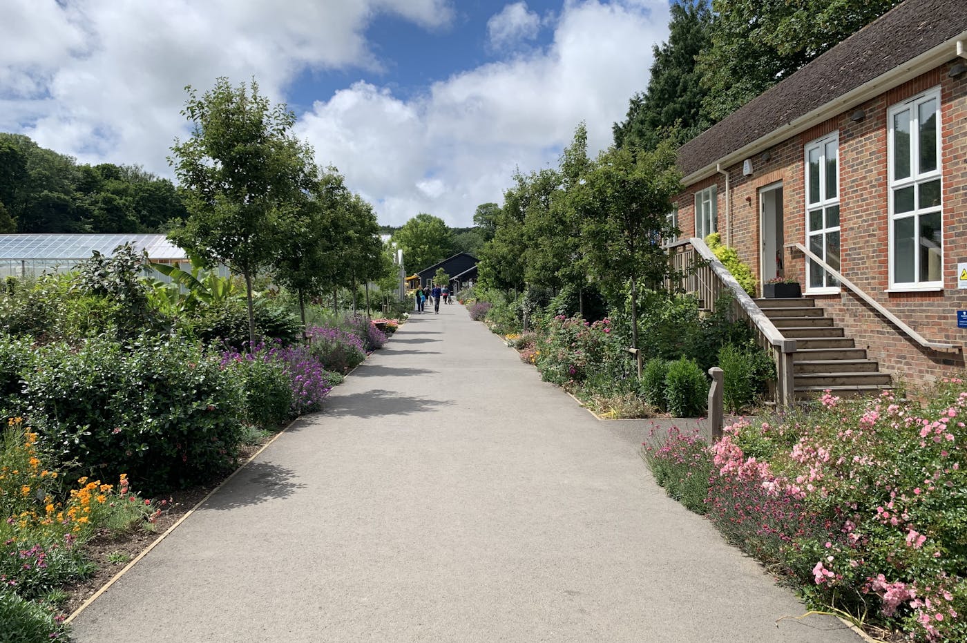 A paved garden path lined with vibrant flower beds leads to a brick building with a glass greenhouse in the background.