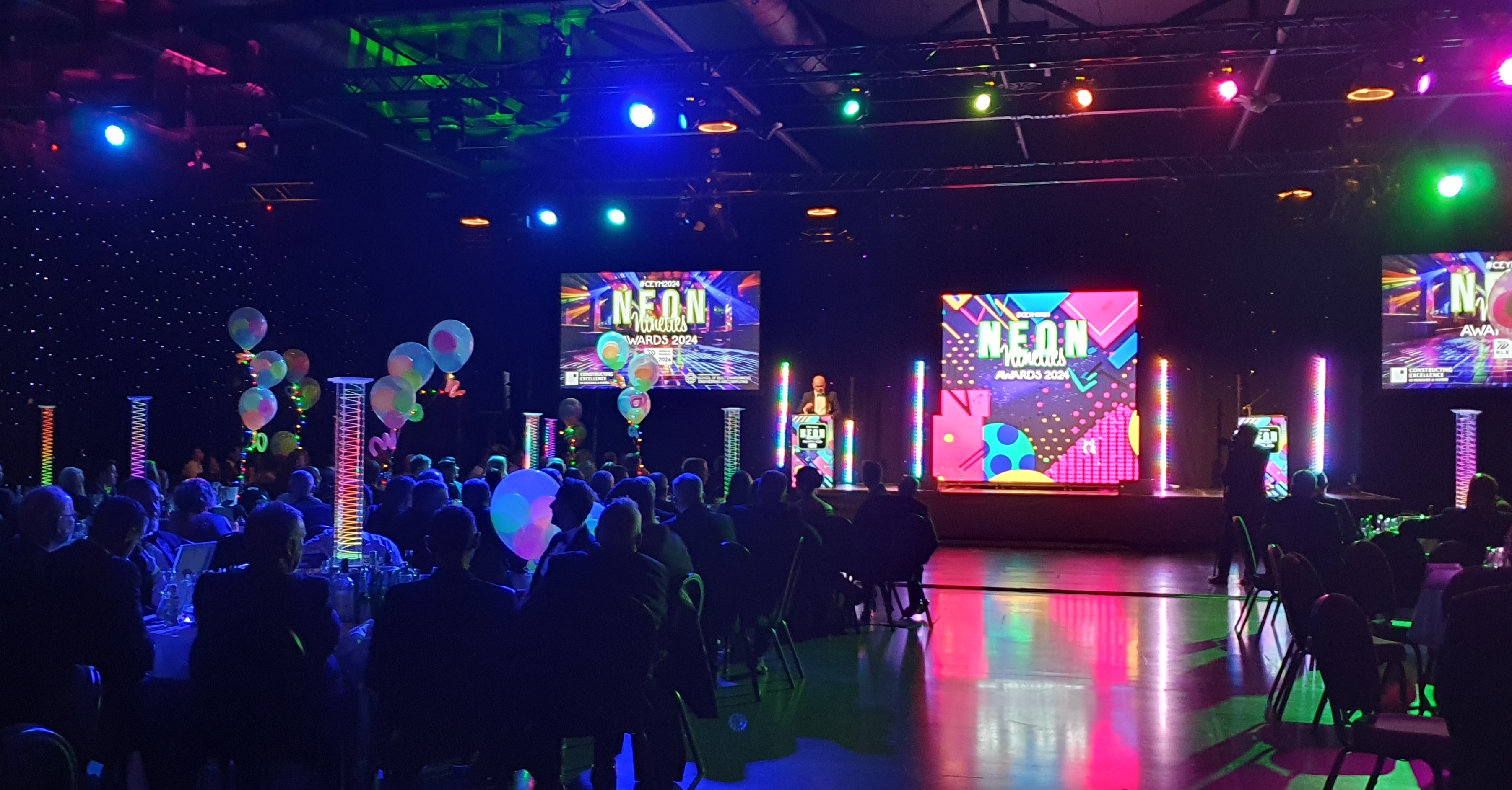 Dark room filled with people sitting around tables with stage and screen at the back of the room