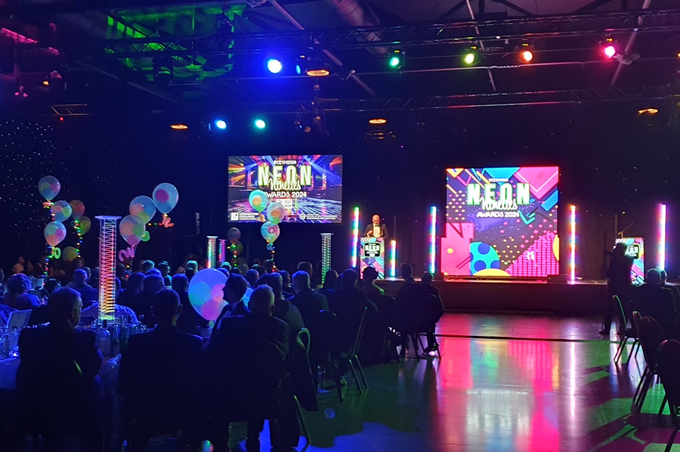 Dark room filled with people sitting around tables with stage and screen at the back of the room