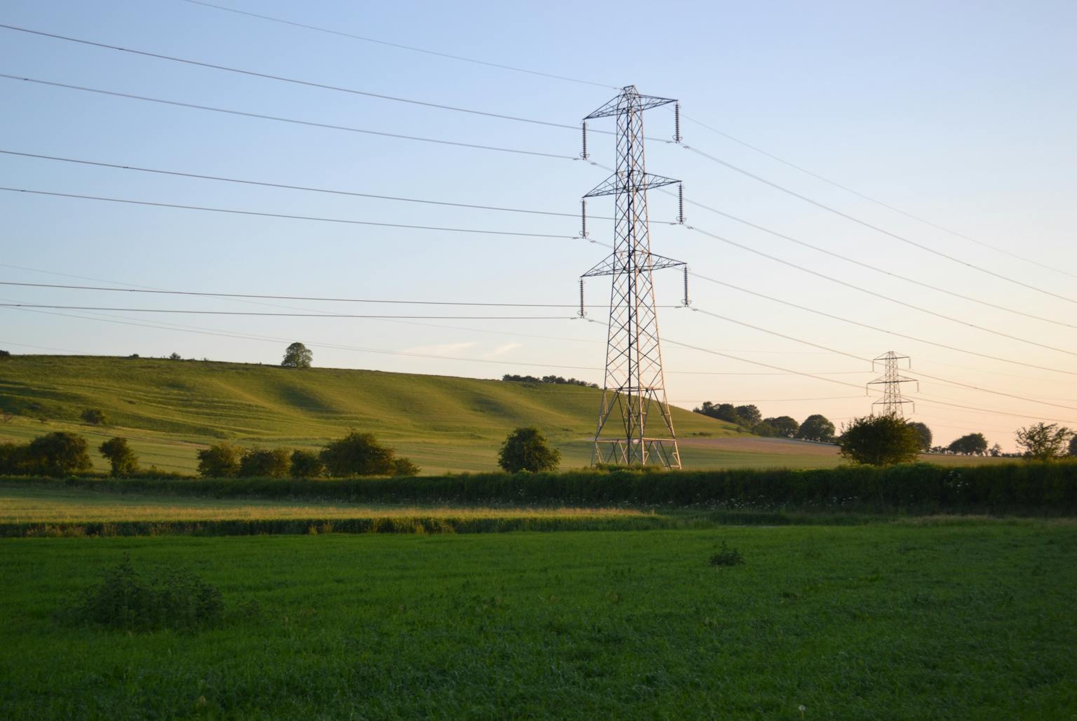 Tall metal electricity pylons stretch across a green field landscape under a clear blue sky.