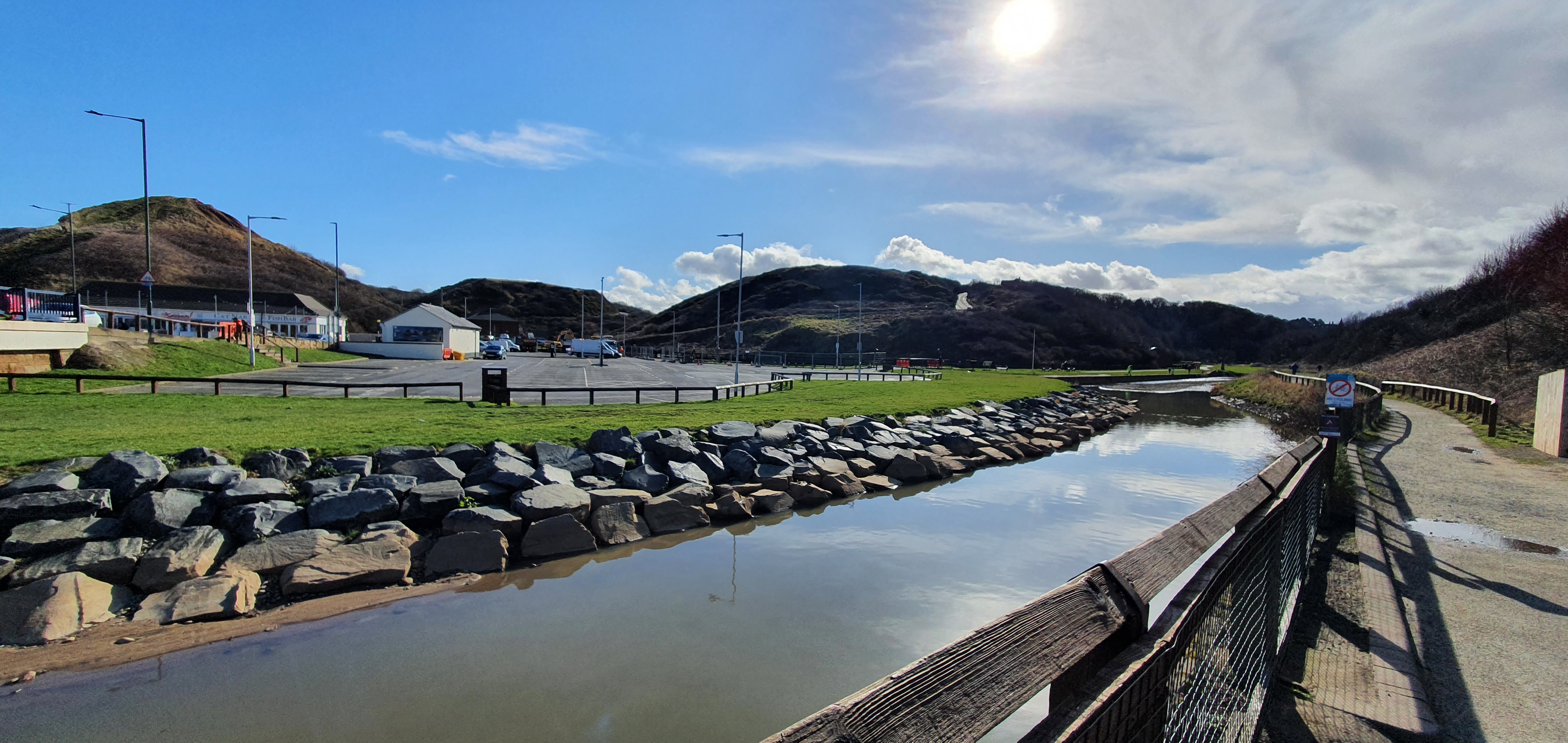 Waterway with stone wall in front of carpark with hills in the background