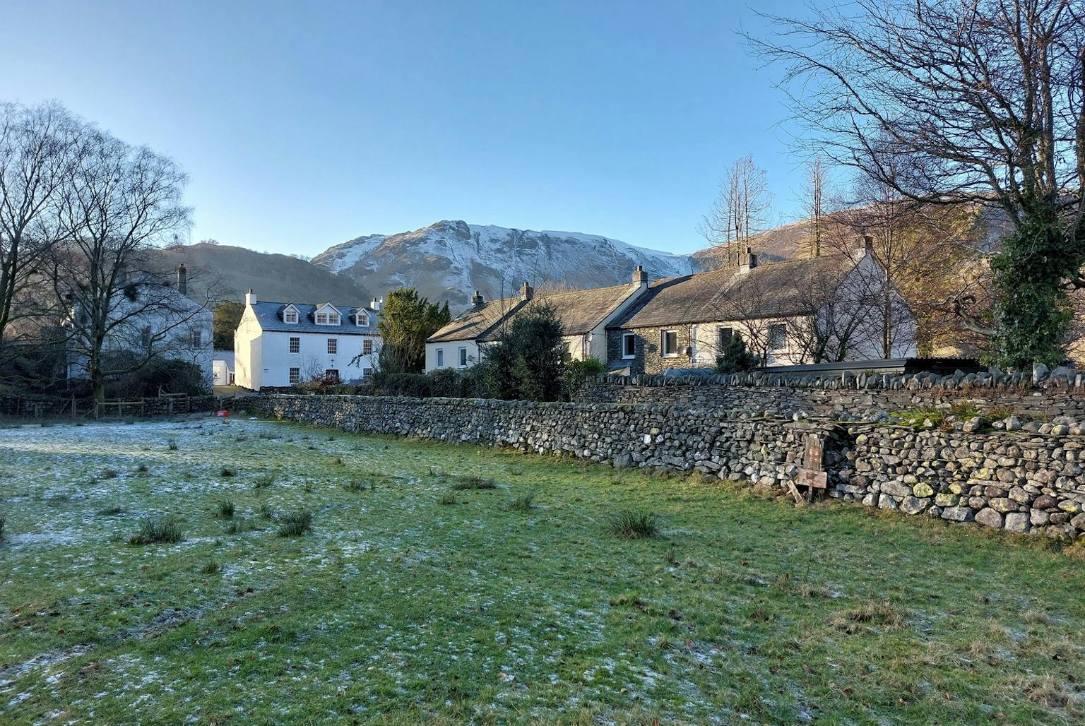 Village comprising buildings positioned behind grass field and brick wall with snow topped mountains in the background