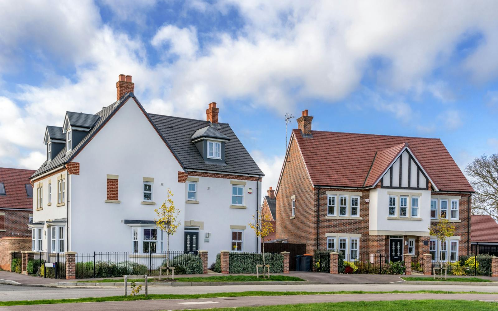 Two modern detached homes with white and red brick exteriors, gabled roofs, and front gardens on a sunny day in a suburban neighbourhood.
