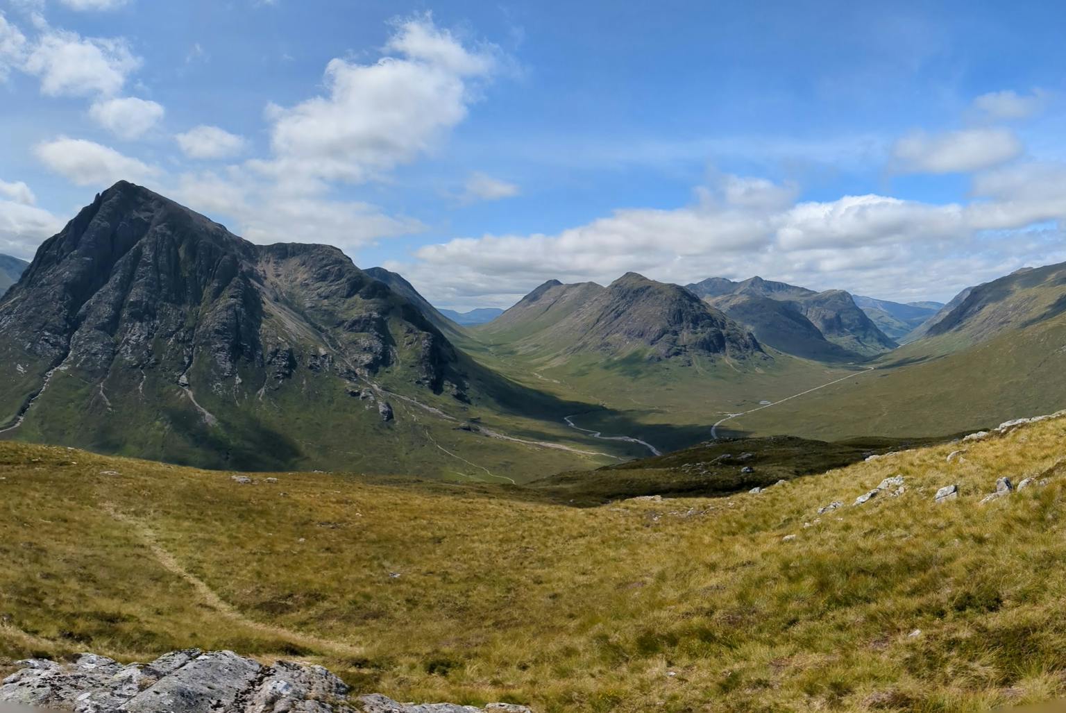 Panoramic view of rolling green hills and mountains under a blue sky with white clouds.