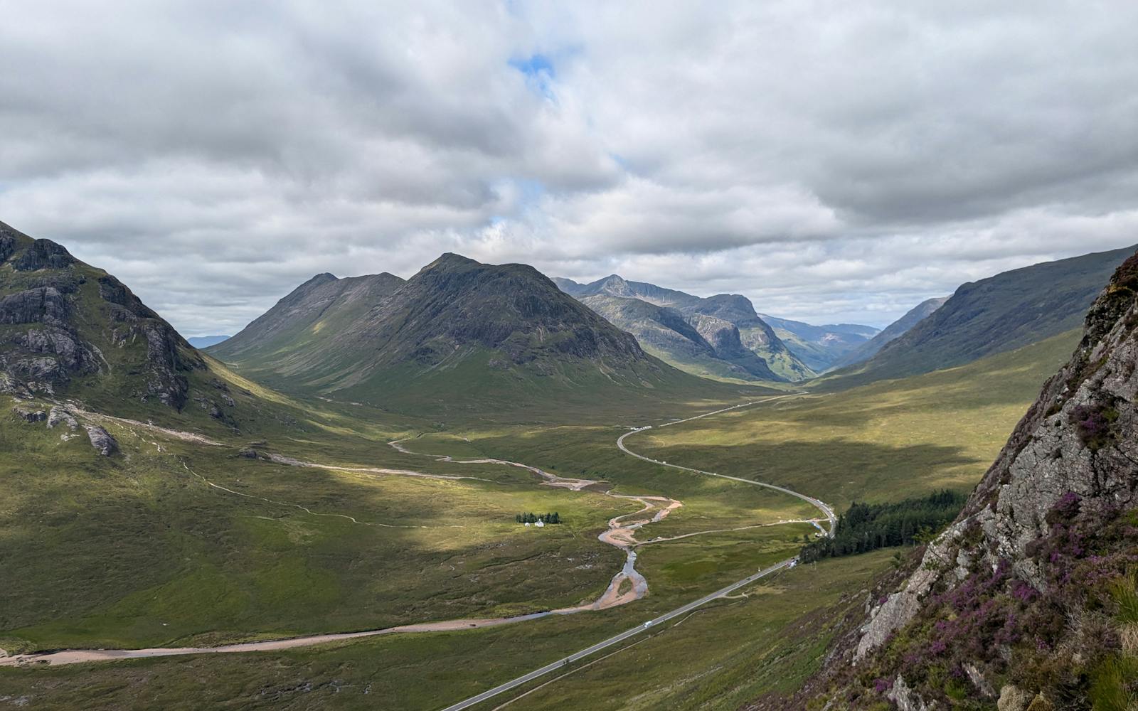 Scenic view of a winding road through a valley surrounded by mountains and hills, with a cloudy sky above.