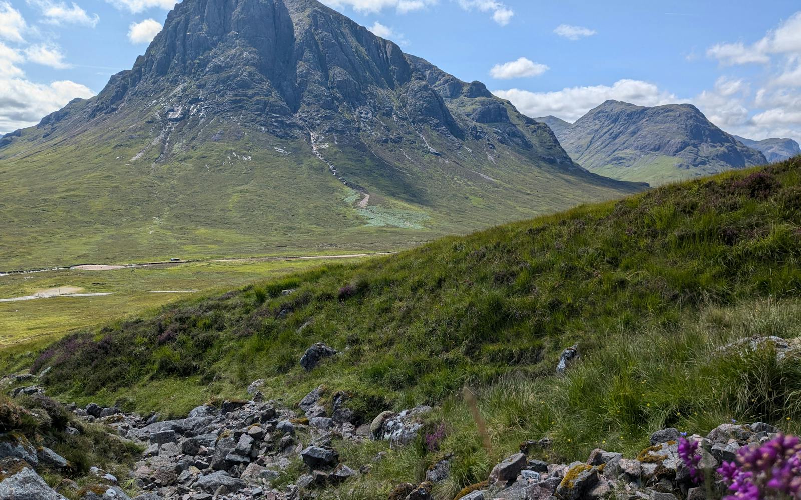 A dramatic mountain peak with a steep, rocky face rises above a valley with green hills and a stream flowing through it. Purple flowers can be seen in the foreground.