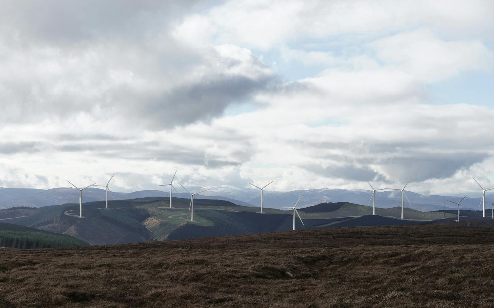 A panoramic view of a wind farm with numerous wind turbines. The turbines are set against a backdrop of rolling hills and a cloudy sky.