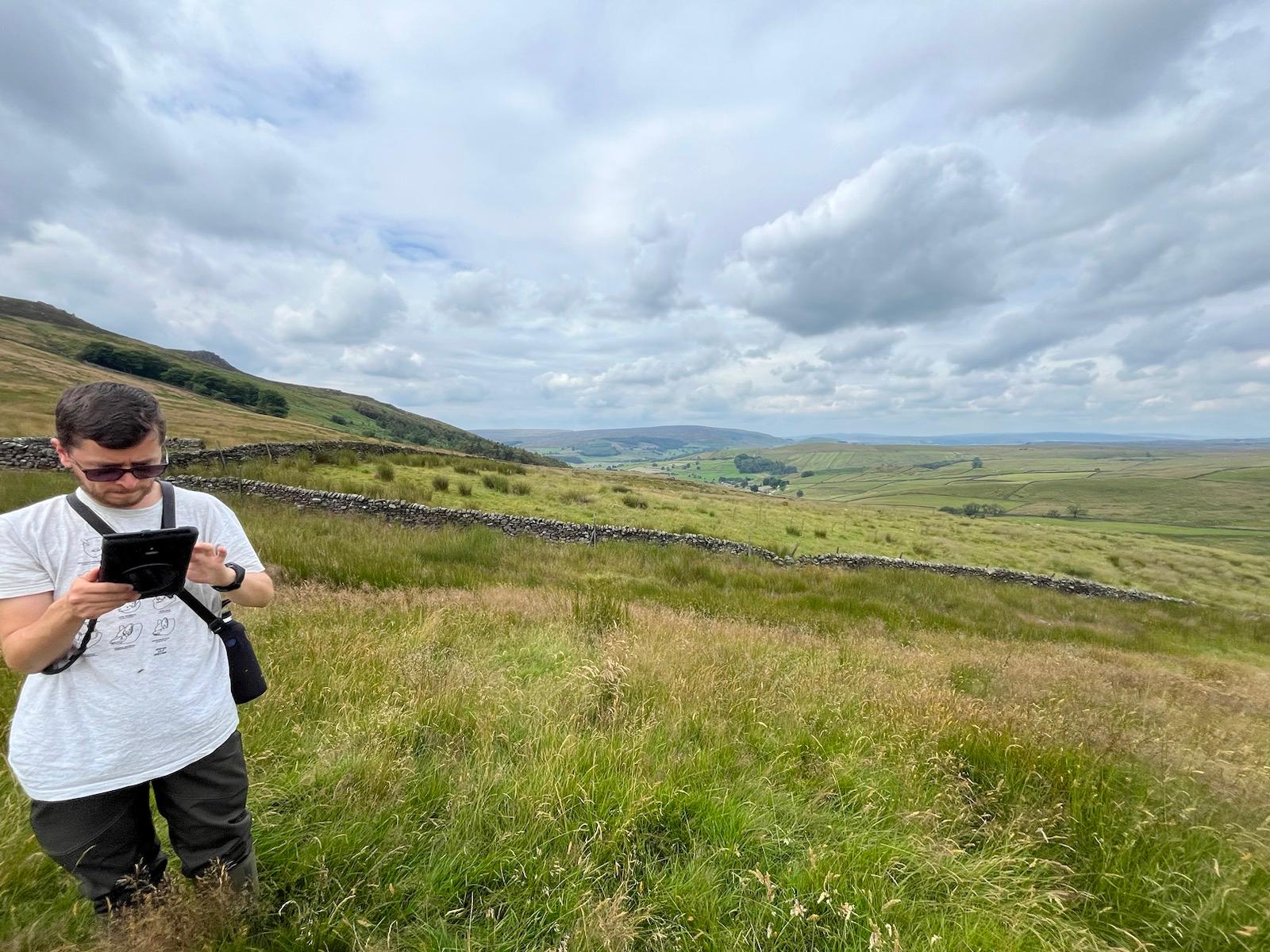 A man wearing sunglasses and a white t-shirt stands in a grassy, rolling landscape, engrossed in using a digital device. Behind him, the vast landscape stretches out with hills, dry stone walls, and fields beneath a cloudy sky. The image captures a peaceful rural environment, highlighting outdoor fieldwork.