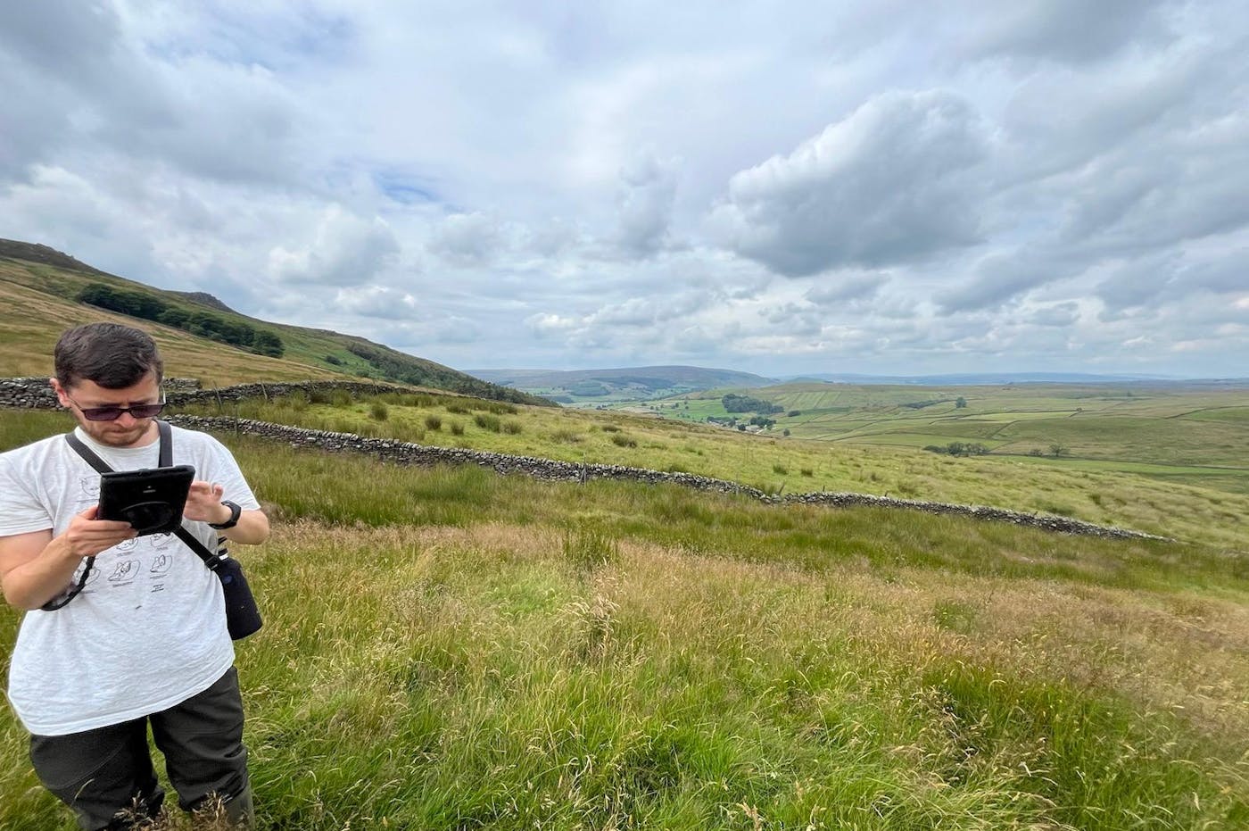 A man wearing sunglasses and a white t-shirt stands in a grassy, rolling landscape, engrossed in using a digital device. Behind him, the vast landscape stretches out with hills, dry stone walls, and fields beneath a cloudy sky. The image captures a peaceful rural environment, highlighting outdoor fieldwork.
