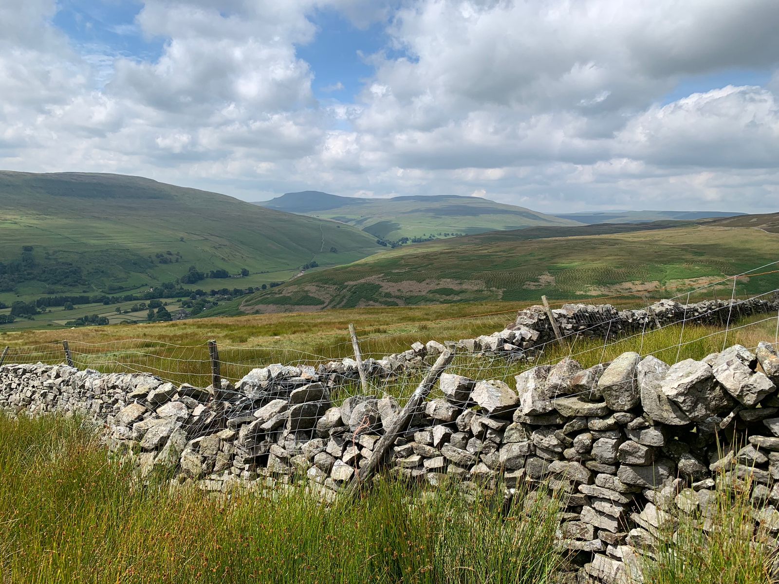 A scenic landscape with a dry stone wall in the foreground. Rolling hills and a distant mountain range are visible under a blue sky with white clouds.