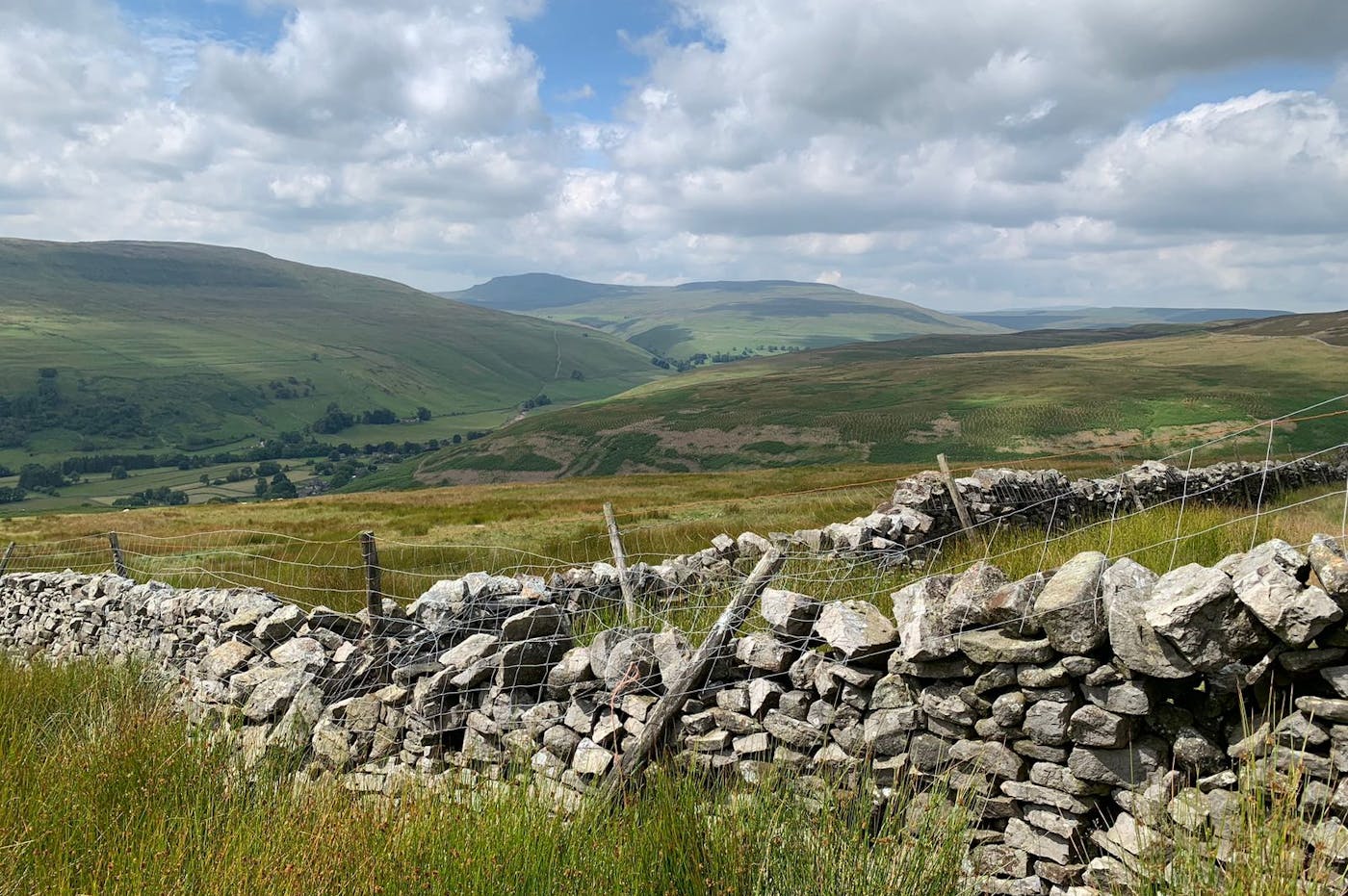 A scenic landscape with a dry stone wall in the foreground. Rolling hills and a distant mountain range are visible under a blue sky with white clouds.
