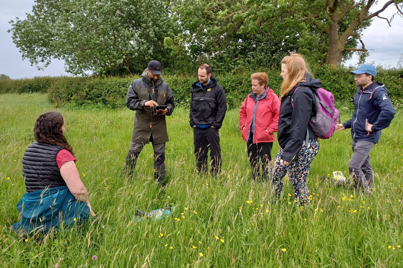 A group of six people stands in a field of tall grass, engaged in conversation. One person sits while the others stand, wearing outdoor gear and jackets. The scene appears to be a meeting or discussion in a natural environment, with trees and hedges in the background on a cloudy day.