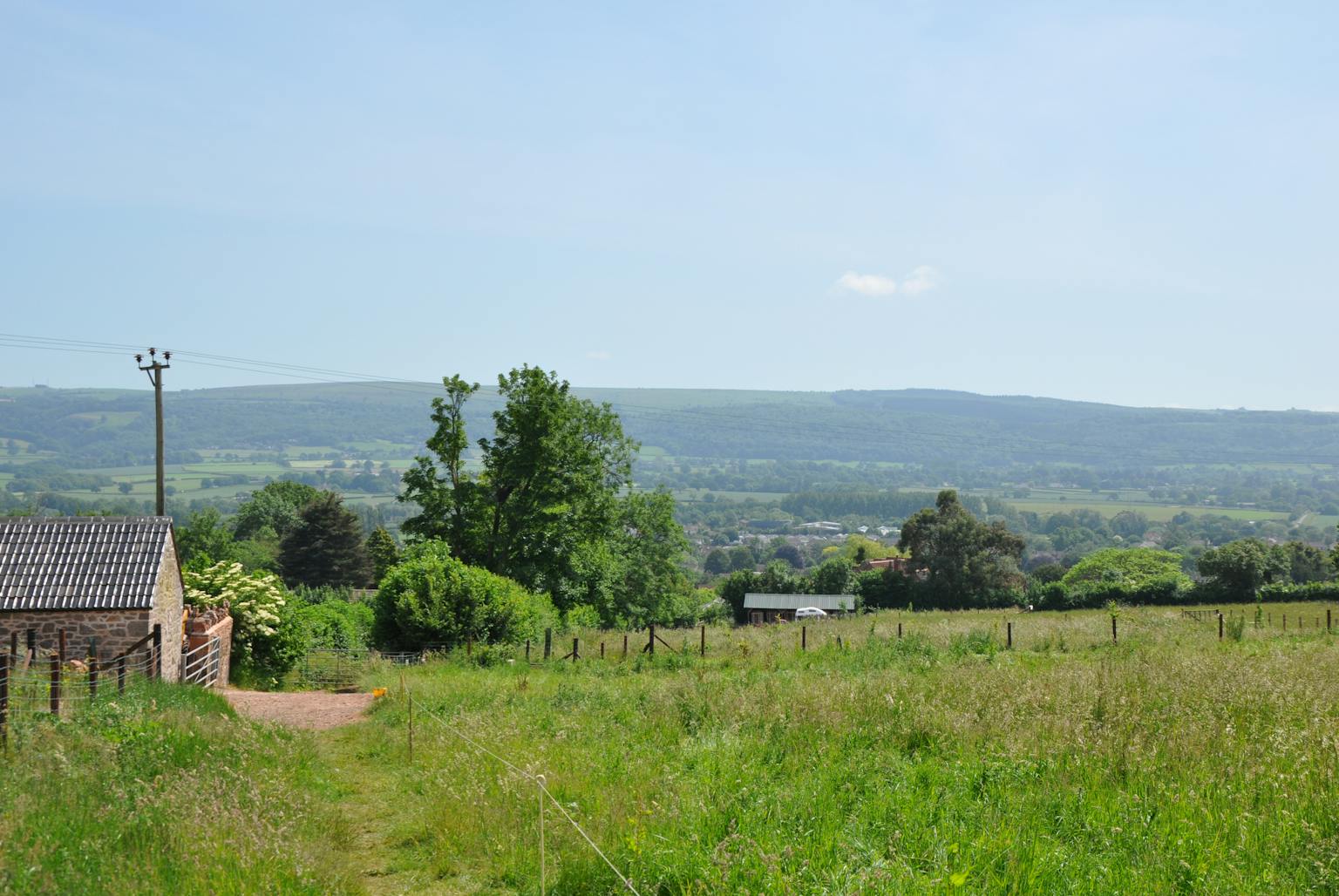 A rural landscape with a field of tall grass, a stone barn, and a fence. In the background, there are rolling hills and a clear blue sky.
