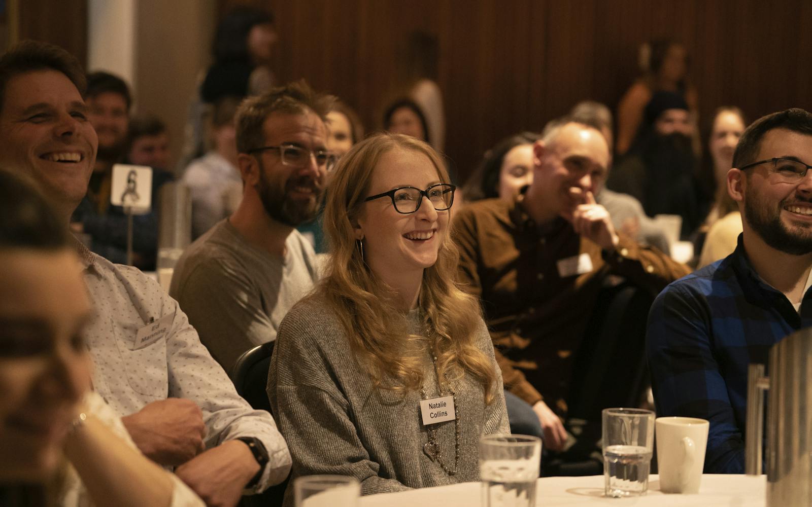 A group of people sit at a table, smiling and engaged in a presentation. The focus is on a young woman with long blonde hair, wearing glasses, a grey jumper, and a name tag reading "Natalie Collins." Glasses of water and a notepad are on the table. The room has a warm, cosy atmosphere with wooden walls in the background.