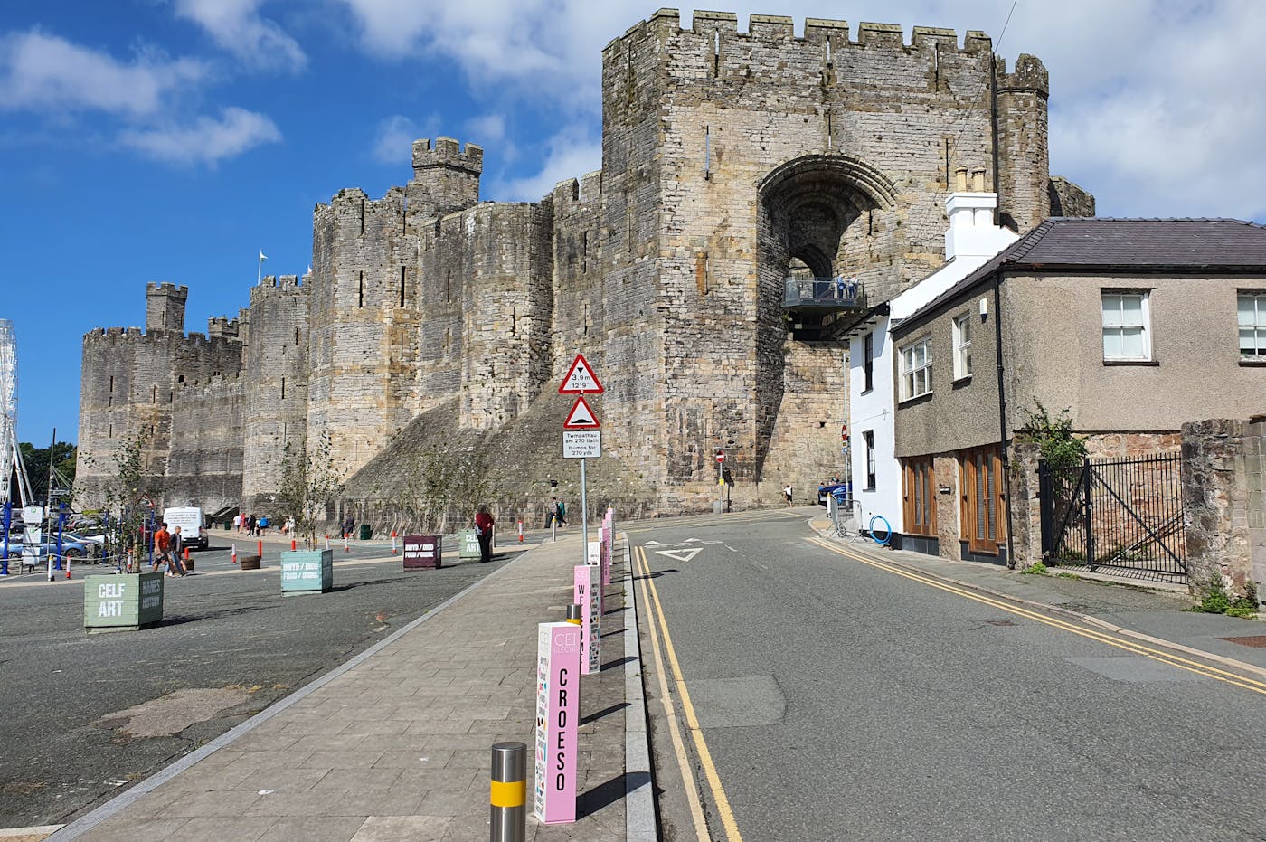 A large castle on a hill with a road leading up to it. There are pink bollards and a sign that says "CROESO."