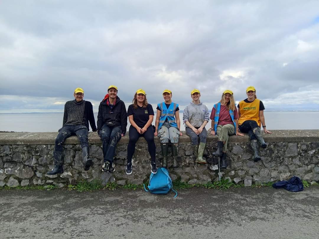 A group of seven volunteers wearing yellow "Marlens" caps sit on a stone wall by the coast. They are smiling, with a cloudy sky and the sea behind them.