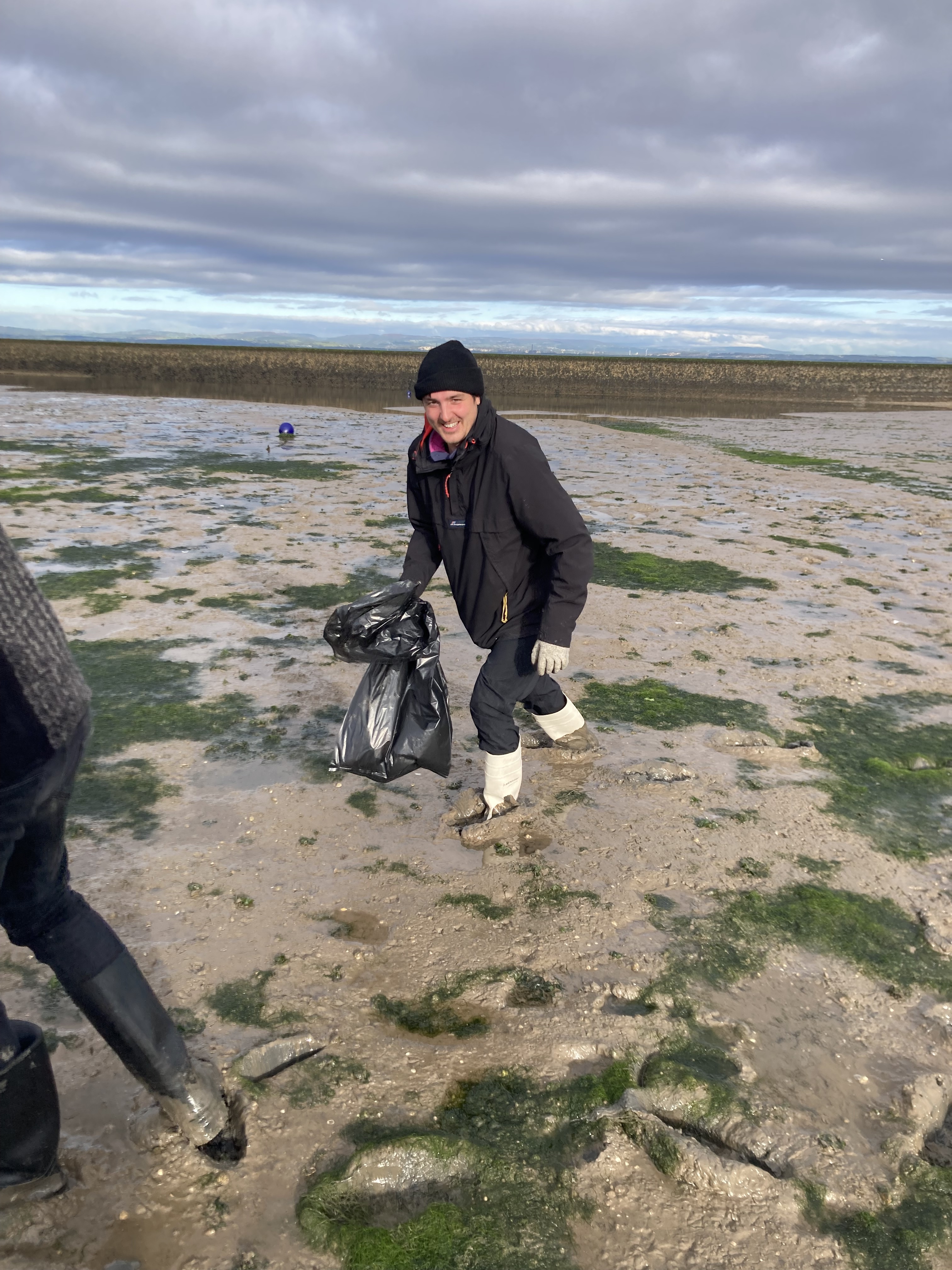 Two individuals stroll along the beach, carrying bags, with waves gently lapping at the shore in the background.