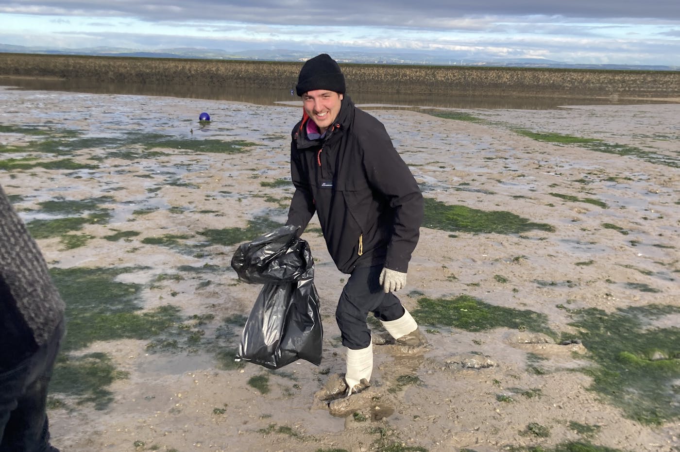 Two individuals stroll along the beach, carrying bags, with waves gently lapping at the shore in the background.