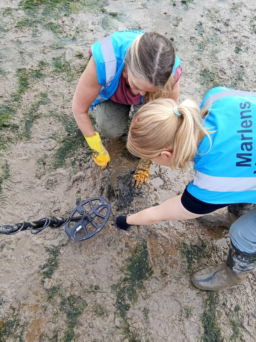Two women in blue vests and yellow gloves are collaboratively working on a metal object in a workshop setting.