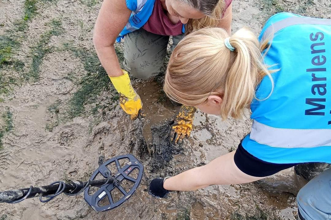 Two women in blue vests and yellow gloves are collaboratively working on a metal object in a workshop setting.