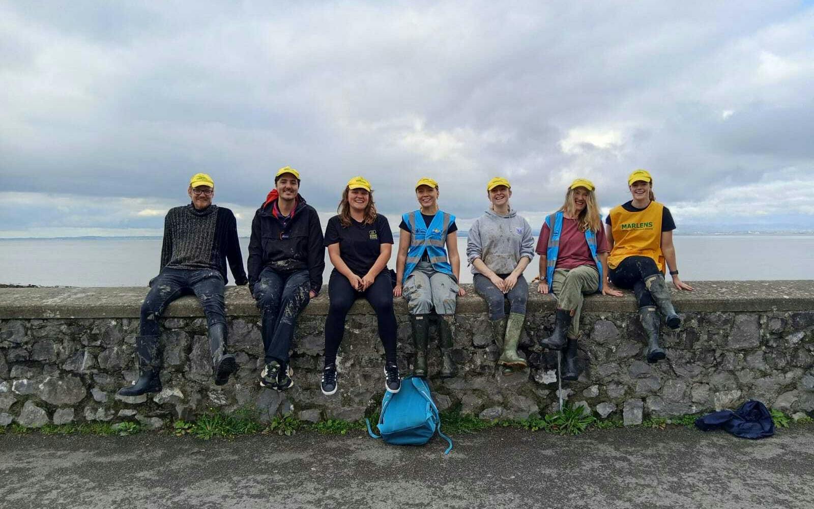 A group of seven volunteers wearing yellow "Marlens" caps sit on a stone wall by the coast. They are smiling, with a cloudy sky and the sea behind them.