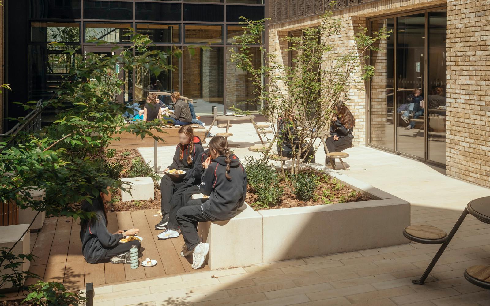 Outdoor courtyard area with students sitting and socialising. Surrounded by modern glass and brick buildings, with trees and plants providing a natural setting. Sunlight casts shadows across the seating and pathways, creating a warm and welcoming atmosphere.