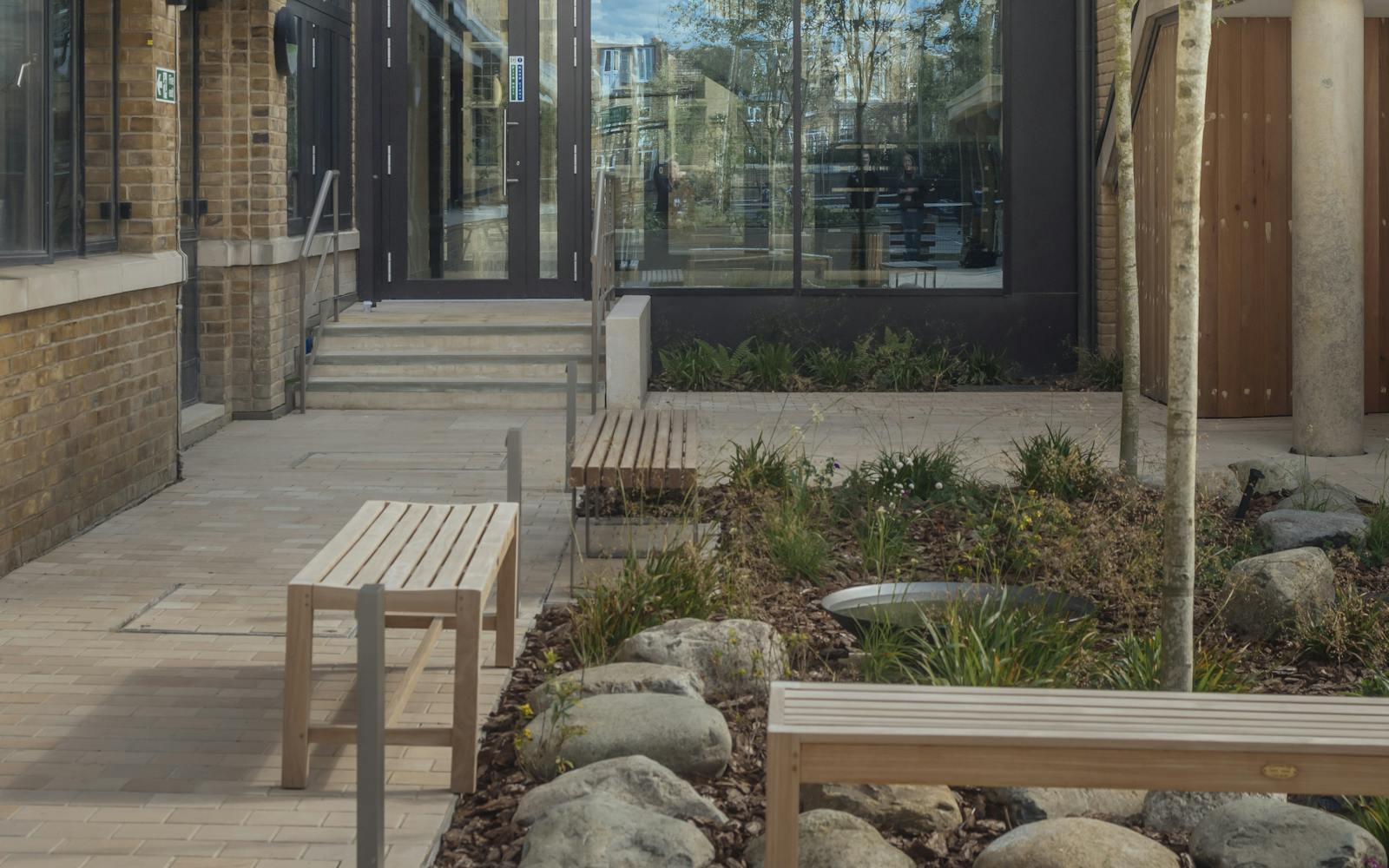 Modern building exterior with large glass windows reflecting the sky and surrounding trees. The courtyard includes wooden benches, plants, and stones, creating a natural, peaceful atmosphere. Brick facades frame the central glass section, where an indoor staircase is visible.