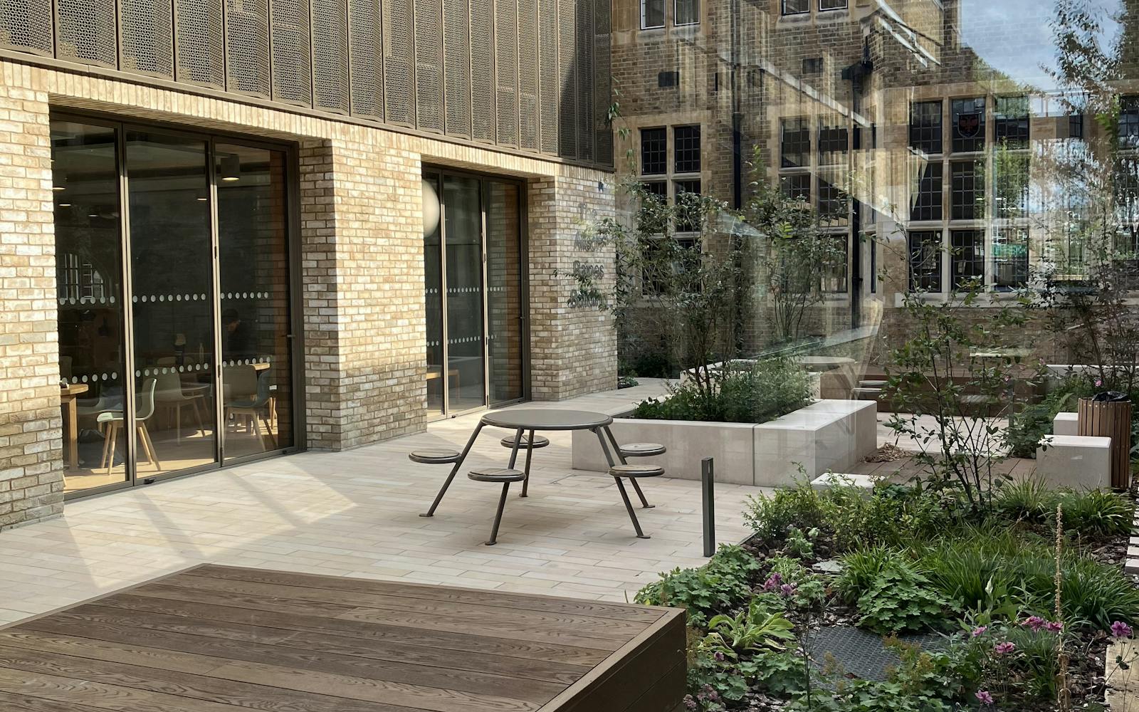 A courtyard with a raised wooden platform and a small table and chairs. The courtyard is surrounded by plants and trees, creating a lush and inviting atmosphere. In the background, a building with large windows reflects the courtyard, creating a sense of depth.