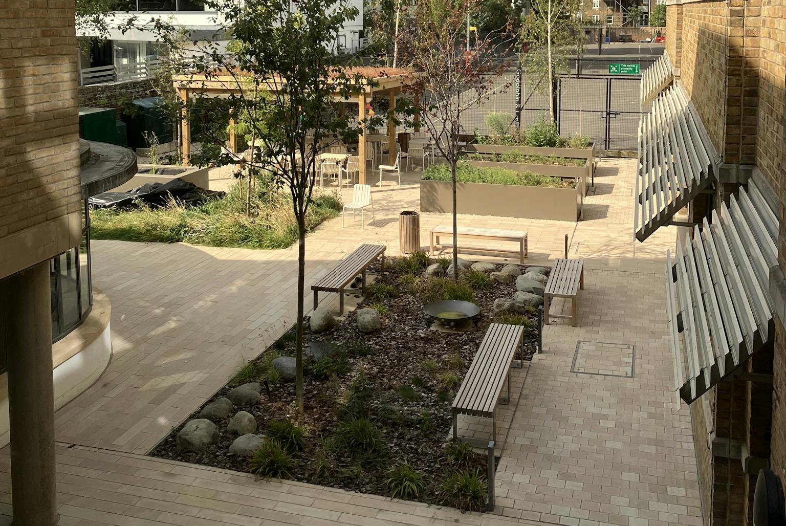 A view of a courtyard with benches, trees, and a small playground in the background. The courtyard is paved with light-colored tiles and has raised planters with plants and small trees. There is a wooden pergola with seating and a small water feature. In the background, you can see a building with the word "FACTORY" on it.