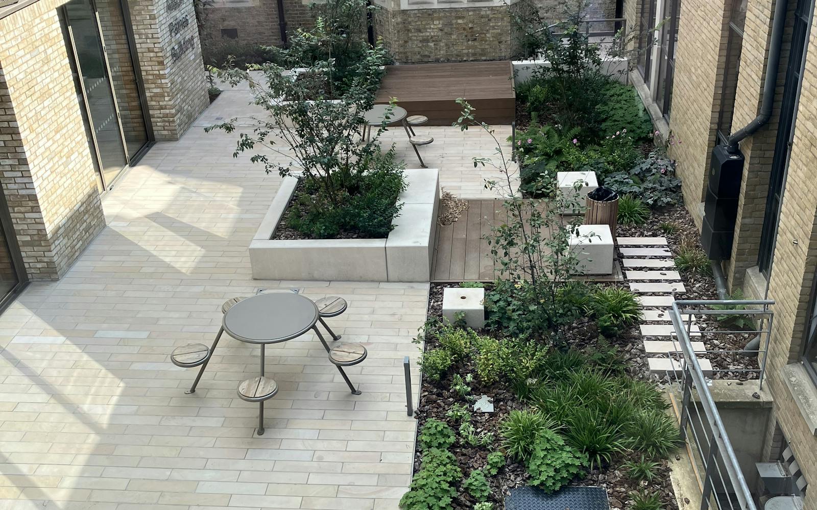 A view of a courtyard with a table and chairs, surrounded by lush greenery and raised planters. The courtyard is paved with light-colored tiles and has wooden decking areas. There are various plants and trees, creating a tranquil and inviting space.