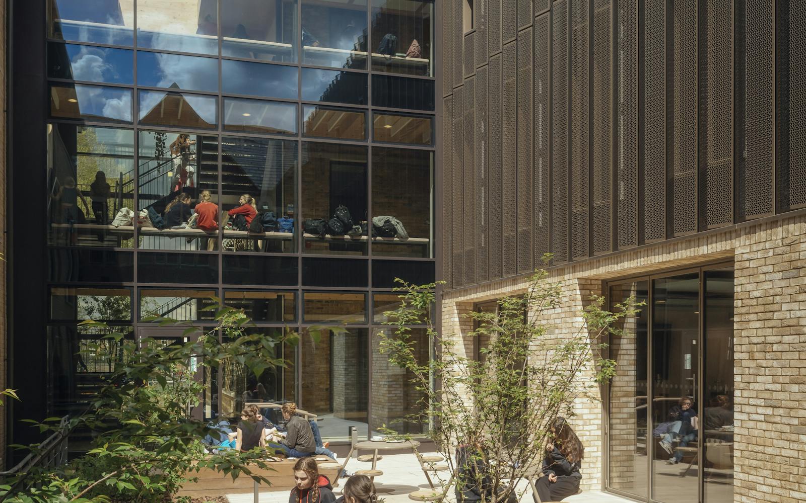 Outdoor courtyard area with students sitting and socialising. Surrounded by modern glass and brick buildings, with trees and plants providing a natural setting. Sunlight casts shadows across the seating and pathways, creating a warm and welcoming atmosphere.